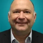 A middle-aged man with a bald head smiles at the camera. He is wearing a checked shirt and a dark jacket. The background is a solid teal colour.
